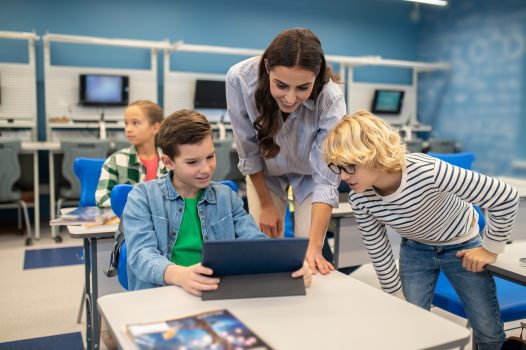 Good job. Smiling woman and boy in glasses looking into tablet of satisfied student sitting at desk in classroom during lesson